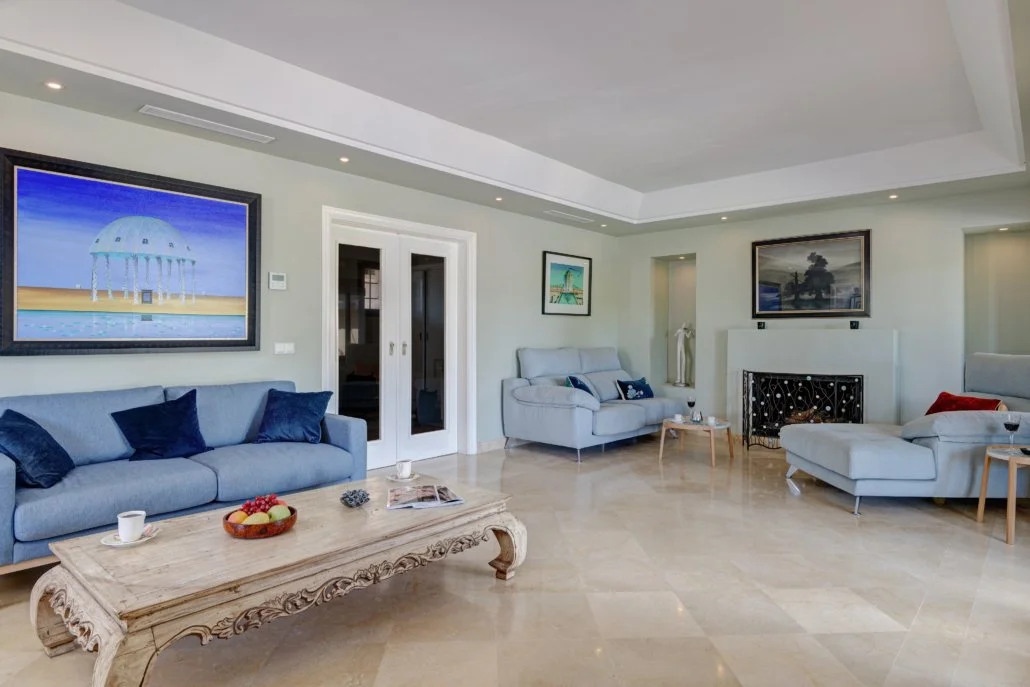 A spacious living room with light-colored walls and marble floors, featuring a large blue sofa with dark blue pillows, a white sofa, and white chaise lounges, all with small side tables. There is a fireplace with a black decorative screen and framed 