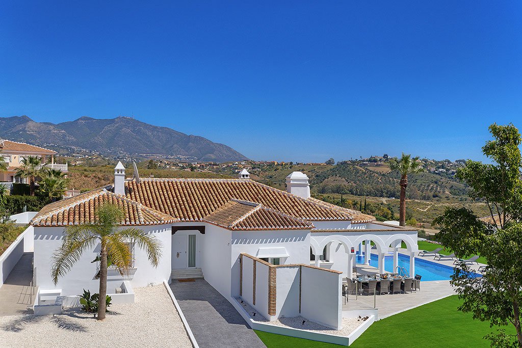 Luxury white villa with terracotta roof, swimming pool, and lush greenery in a scenic landscape with mountains in the background.