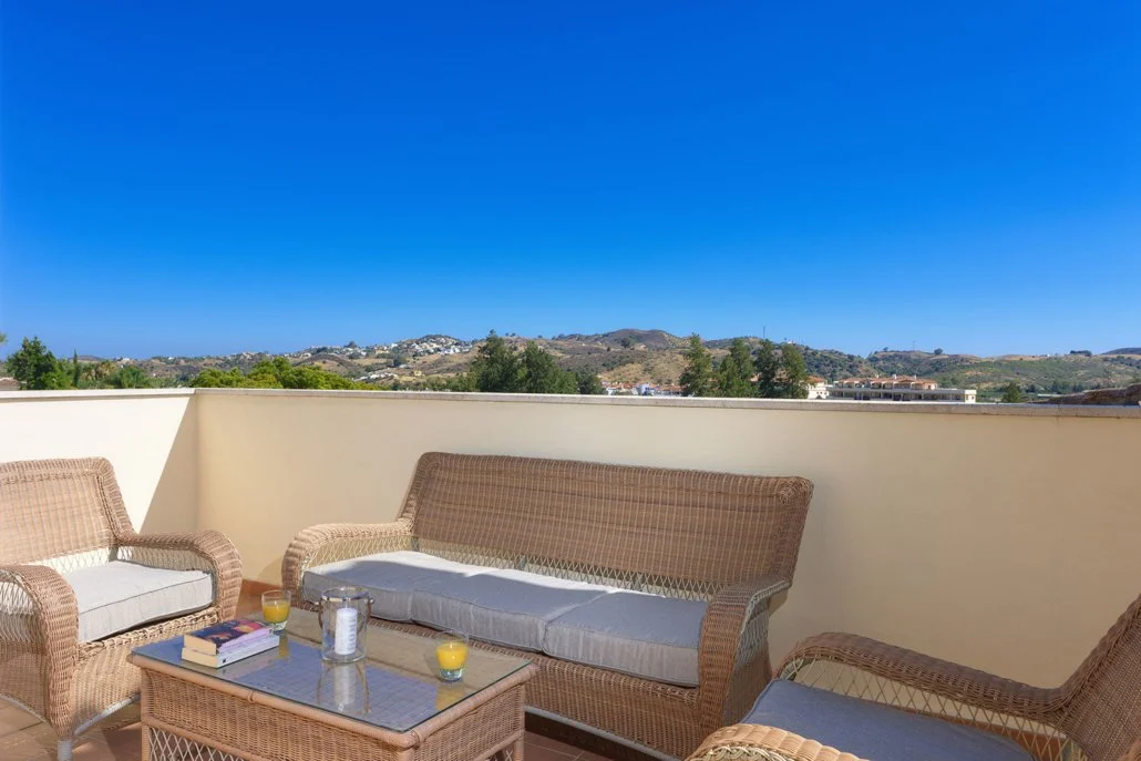 Outdoor balcony with wicker furniture, a table, orange juice, books, and a clear blue sky with distant hills.