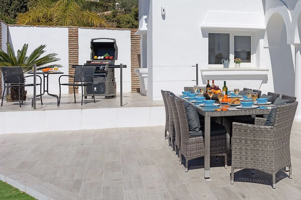 Outdoor patio with a dining table set for a meal, including blue bowls, wine bottles, and glasses, and a grilling area with chairs and a grill in the background.