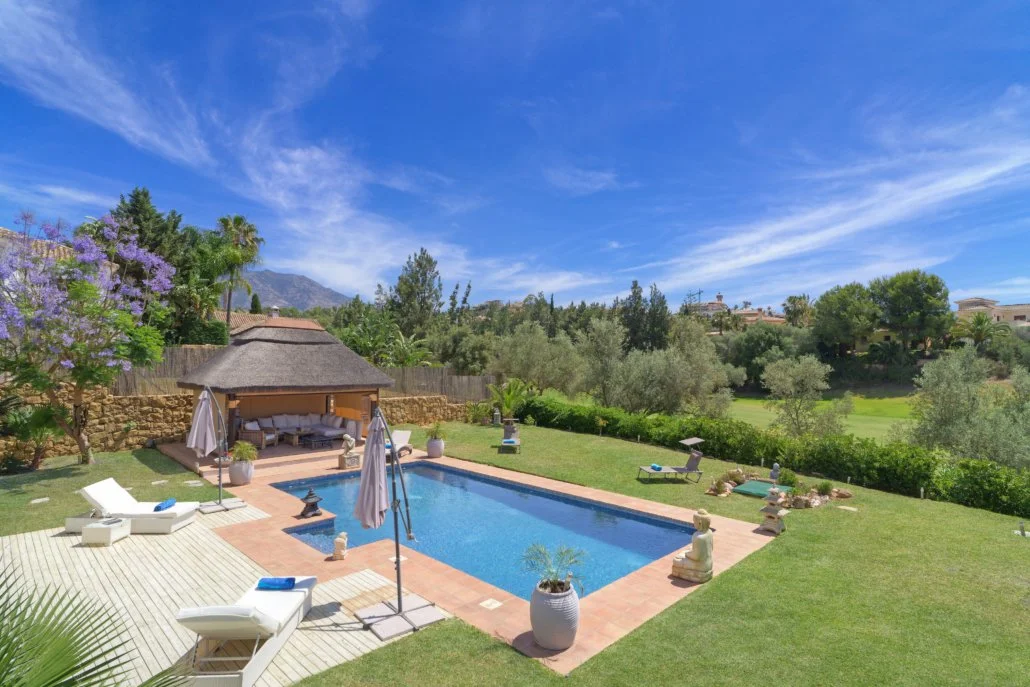 A backyard with a rectangular swimming pool, surrounded by lawn chairs, umbrellas, and lush trees with mountains in the background.