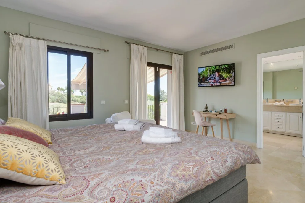 Hotel room with a large bed, two windows with curtains, a small desk with a chair, a wall-mounted TV, and a bathroom visible through an open door.