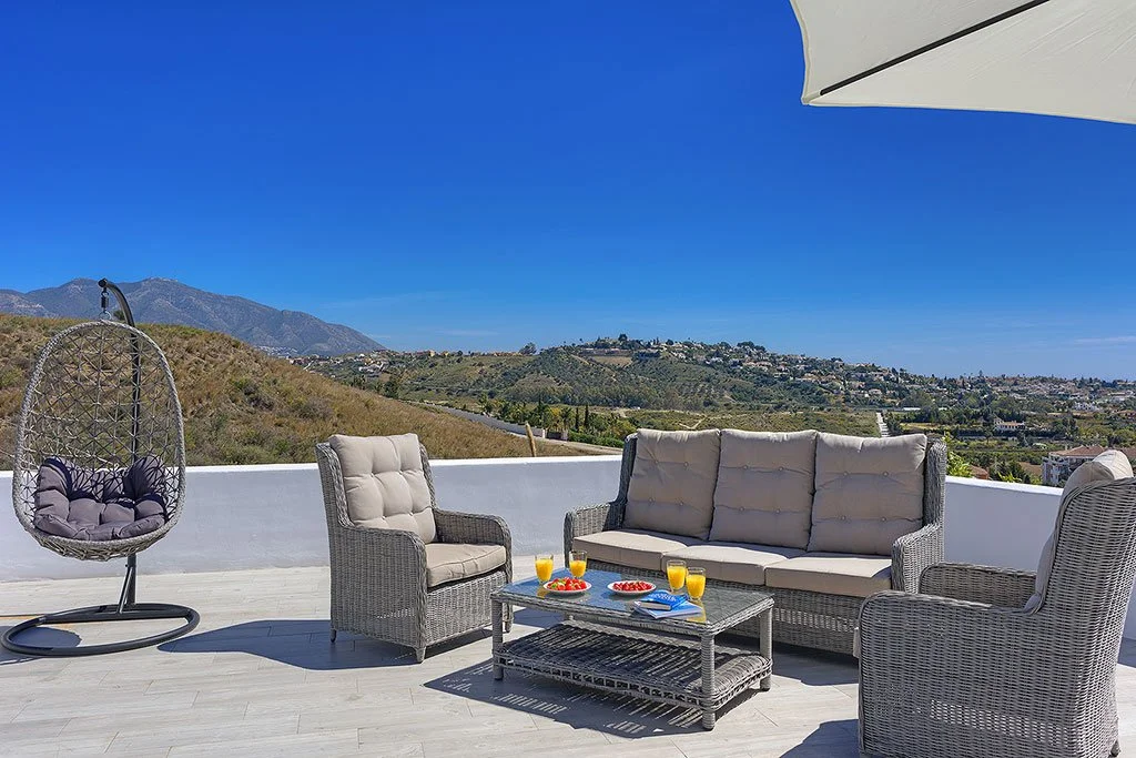 Outdoor patio with wicker furniture, cushioned chairs, sofa, a hanging egg chair, a table with breakfast items, and an umbrella overlooking hills and mountains under a clear blue sky.