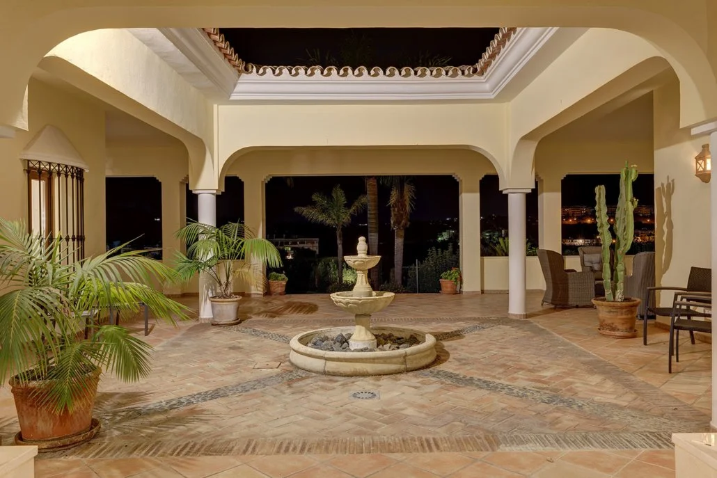 Night view of a spacious covered patio with a decorative fountain at the center, potted plants, wicker and metal chairs, and arches framing a cityscape with palm trees and lights in the background.
