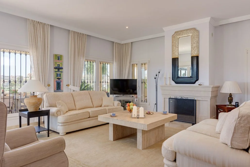 Living room with beige sofas, a fireplace with a mirror above, a TV, side tables with lamps, a coffee table, and large windows with curtains.