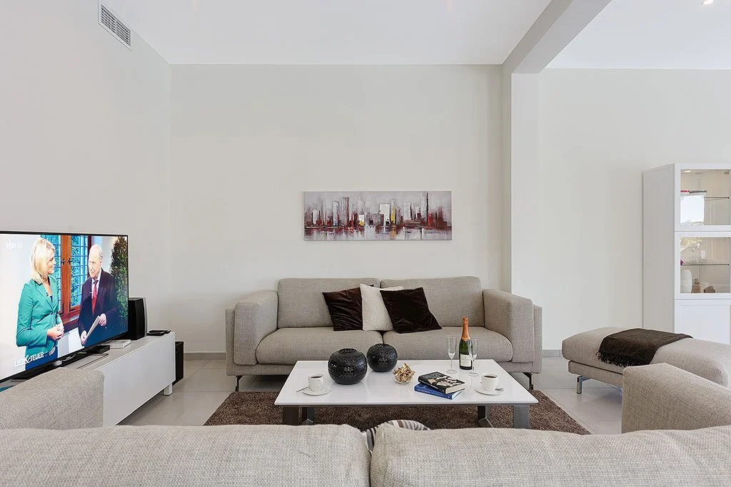 A modern living room with a beige sofa, a coffee table with vases, a TV showing two people talking, and minimalistic wall art.