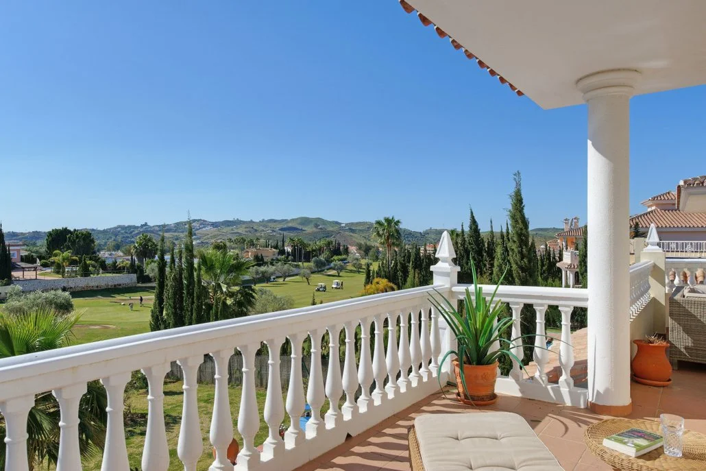 View from a balcony overlooking a golf course with trees and distant hills, sunny day, no people, and patio furniture.
