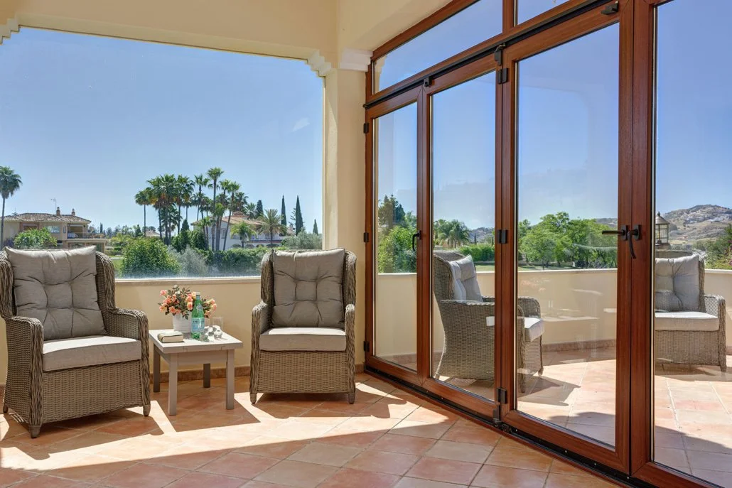 Sunlit enclosed patio with wicker chairs, a side table with flowers, and large glass doors leading to an outdoor area with trees and houses in the distance.