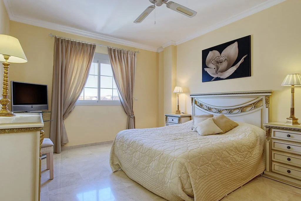 A bedroom with a large bed that has a beige quilt and pillows, cream-colored walls, a window with beige curtains, nightstands with lamps on each side, a flat-screen TV on a dresser, and a wall art featuring a flower.