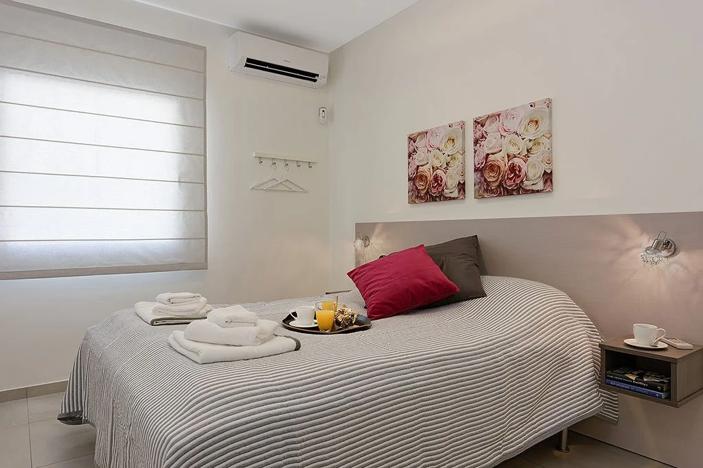 A neatly made bed with gray and white striped bedding, two pillows (one gray, one red), with towels and breakfast items on top, in a minimalistic bedroom with white walls, floral artwork, and a window with a white shade.