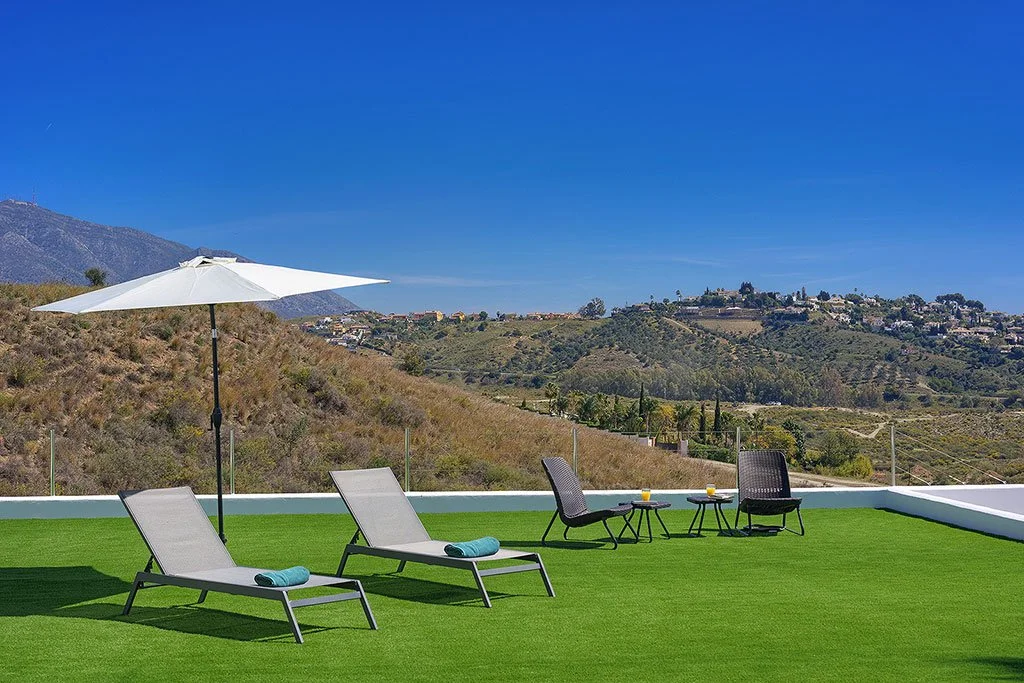 Rooftop patio with two lounge chairs, an umbrella, and two small tables with drinks, overlooking a hillside landscape.