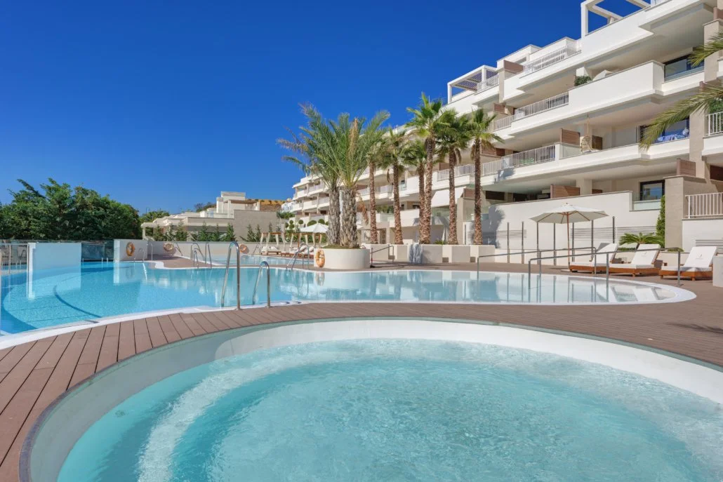 Hotel pool area with a hot tub, lounge chairs, umbrellas, tall palm trees, and a modern white building with multiple balconies under a clear blue sky.