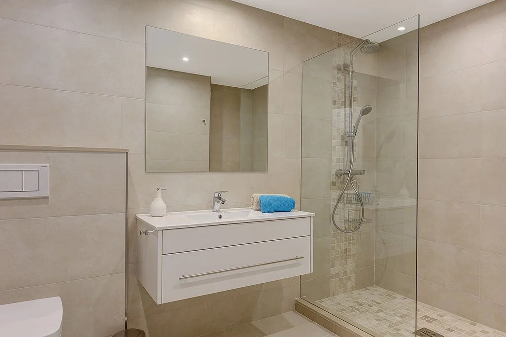 Modern bathroom with a walk-in shower, white vanity with a mirror, beige tiles, and a blue towel.