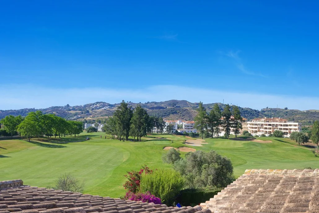 View of a golf course with green fairways and sand traps, surrounded by trees, with residential buildings and hills in the background under a clear blue sky.
