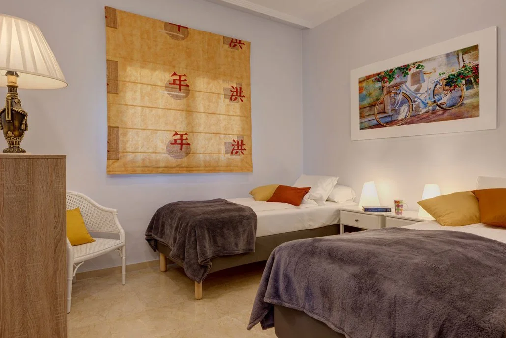 A cozy bedroom with two beds, each with white sheets and brown blankets, and yellow and brown pillows. There are two bedside lamps on white nightstands, a white chair with a yellow pillow on it, and two large framed pictures on the walls—one of a bic