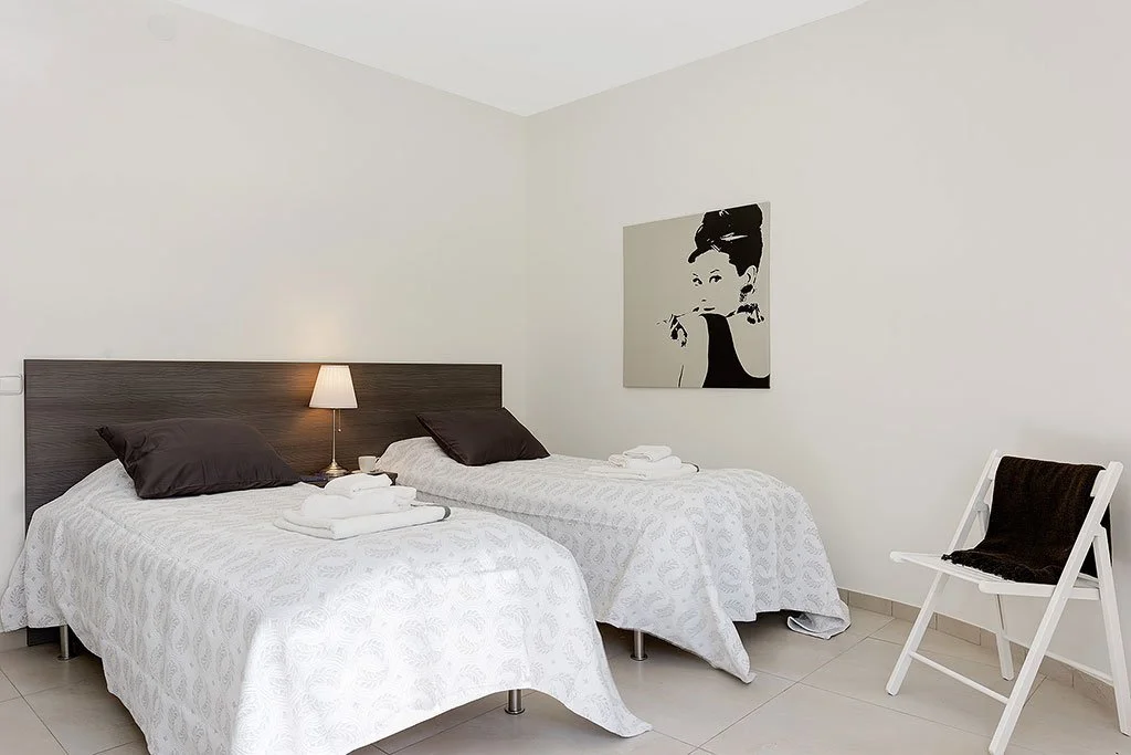 A hotel room with two beds, a nightstand with a lamp, and a piece of artwork of a woman on the wall.