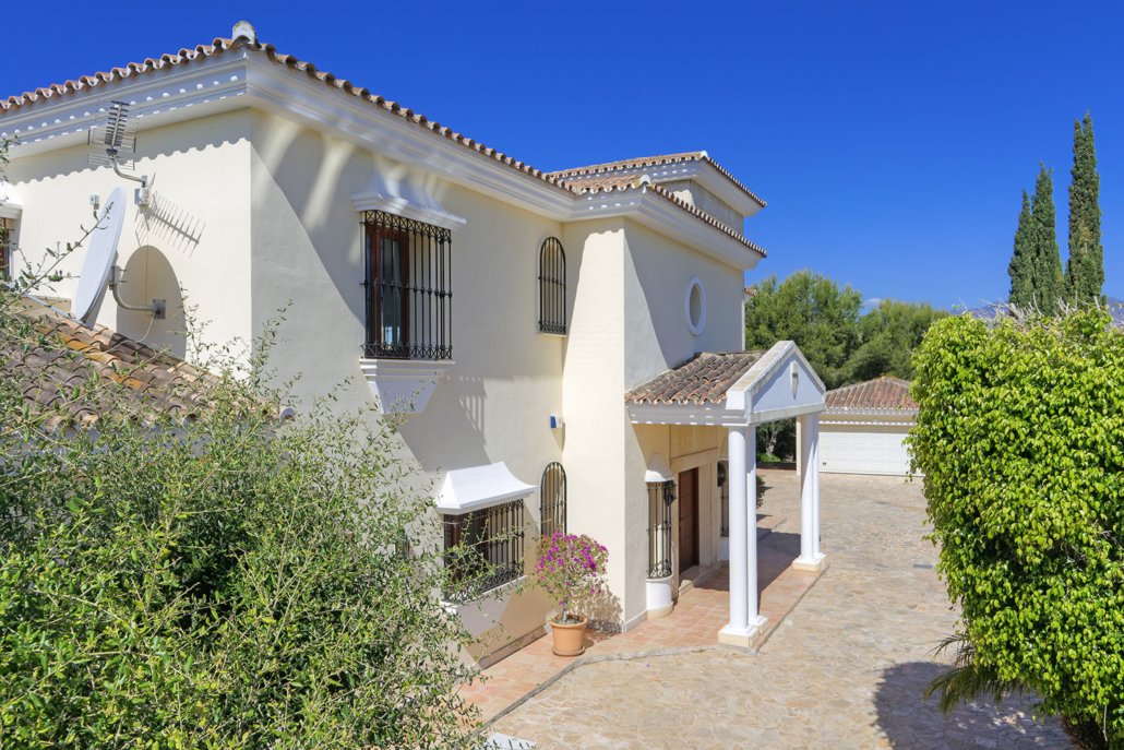 A white Mediterranean-style house with a tiled roof, black wrought iron window bars, and an entrance with white columns and a gabled roof, surrounded by shrubs and trees under a clear blue sky.