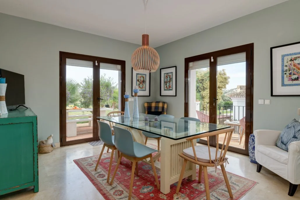 Dining area with a glass-top table, six chairs, and a vintage rug. French doors leading to a balcony with outdoor seating, framed artwork on light green walls, and a mix of modern and vintage decor.