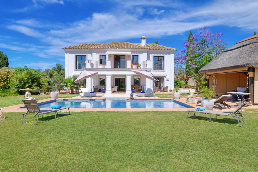 A luxurious backyard with a swimming pool, lounge chairs, umbrellas, and a white two-story house with balconies under a blue sky.