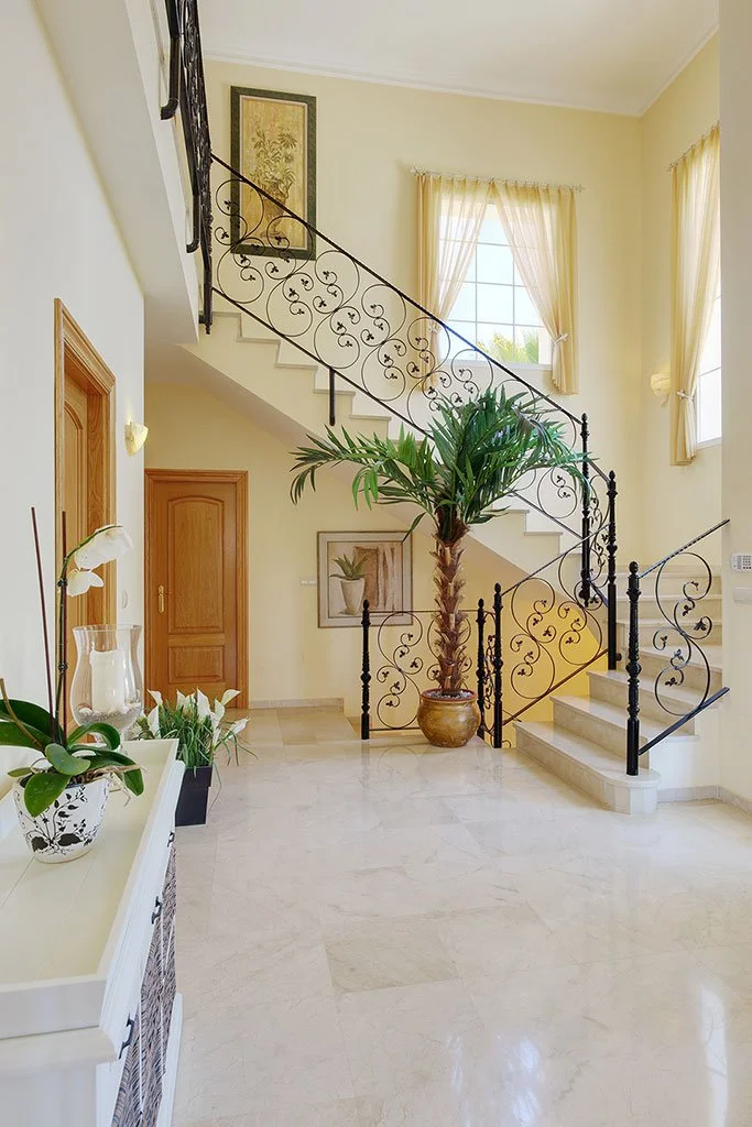 Bright entryway with marble flooring, a large potted palm, decorative iron staircase railing, framed artwork, and light yellow walls with windows and curtains.