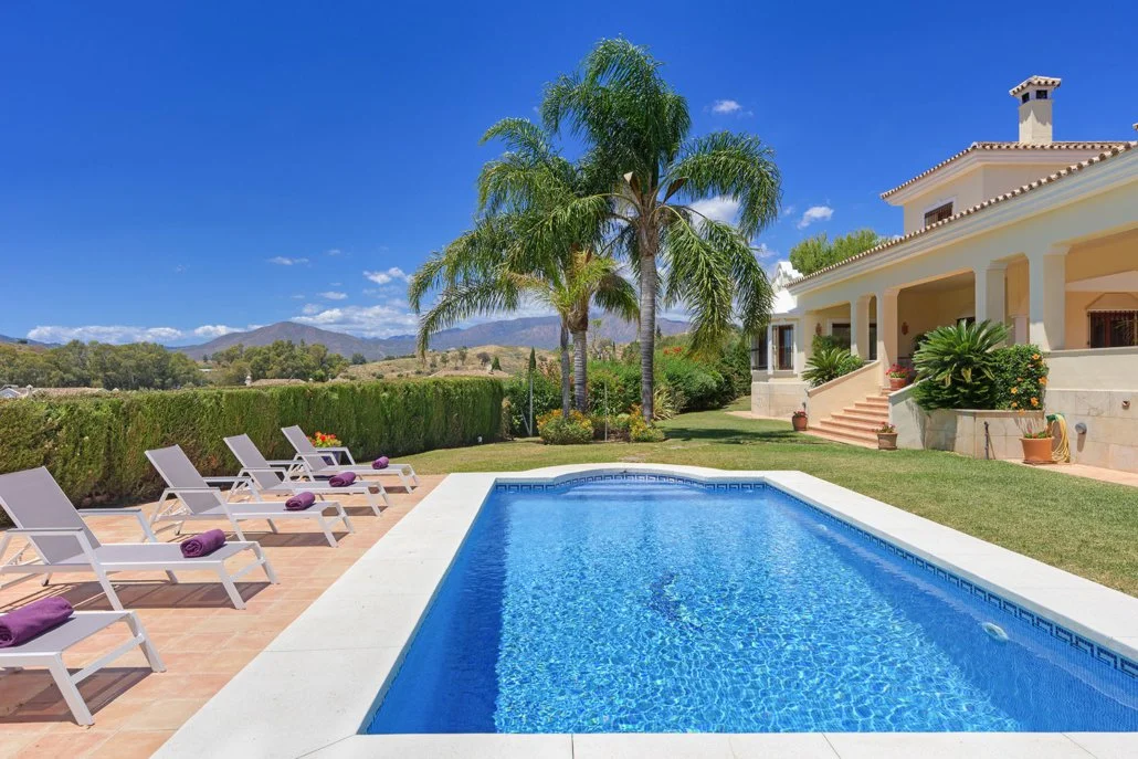 Luxury house with outdoor pool, lounge chairs, palm trees, and scenic mountain view under a blue sky.