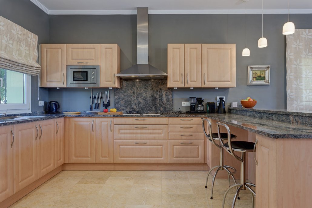 Modern kitchen with light wood cabinets, granite countertops, and gray walls. Contains a window, microwave, range hood, and various small appliances. Bar stools are at the counter, with pendant lights overhead.