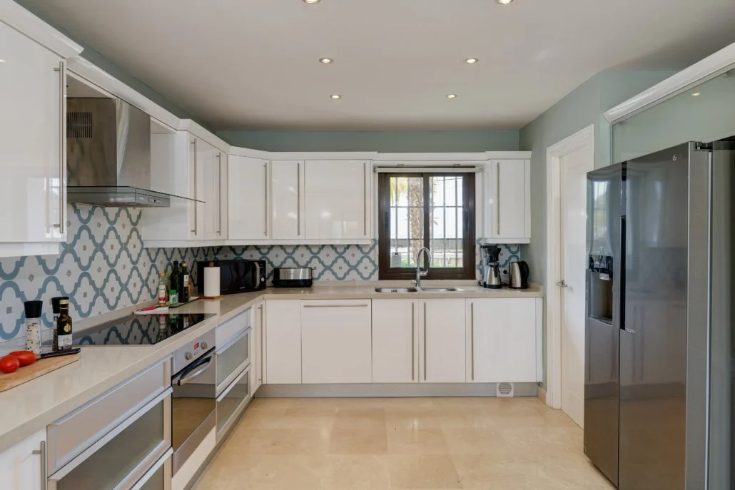 Modern kitchen with white cabinets, patterned backsplash, and stainless steel appliances, including a refrigerator, oven, and microwave. There are small appliances and condiments on the countertops, and a window above the sink.