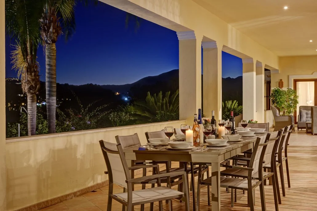 Outdoor dining table set with wine glasses, bottles, and candles, overlooking a view of mountains and palm trees during evening.