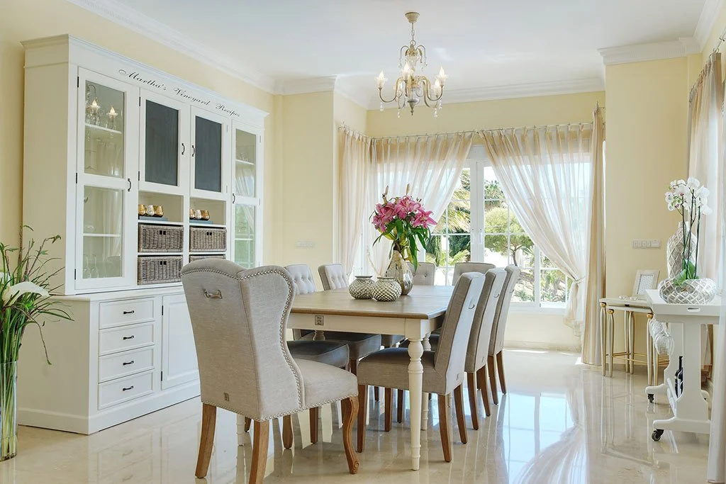 Bright dining room with beige walls, a chandelier, a white dining table with eight chairs, a white cabinet, and windows with sheer curtains, decorated with flowers.