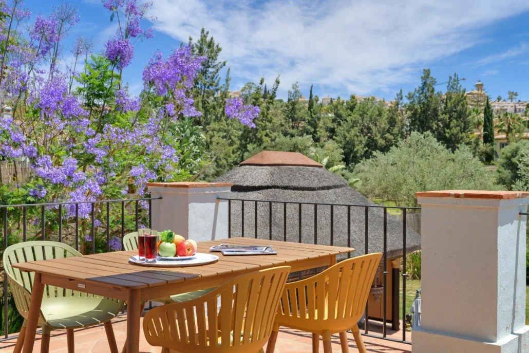 Outdoor patio scene with wooden table and chairs, purple flowering bushes, green trees, blue sky, and rooftops in the background.