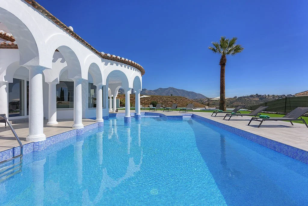 A swimming pool in front of a white building with arches, surrounded by lounge chairs, with mountains and a clear blue sky in the background.
