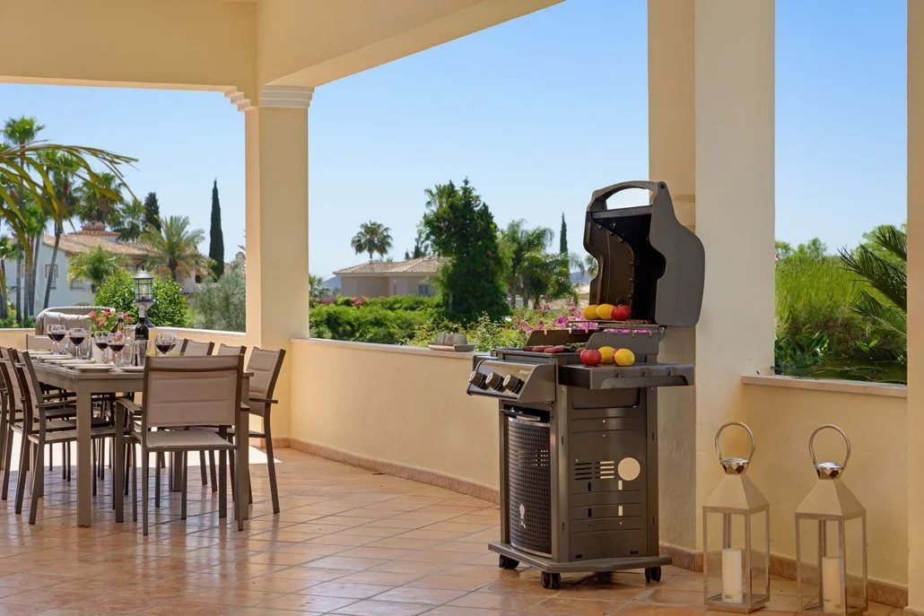 An outdoor patio with a dining table set for wine and a barbecue grill, overlooking a lush garden with palm trees under a bright blue sky.
