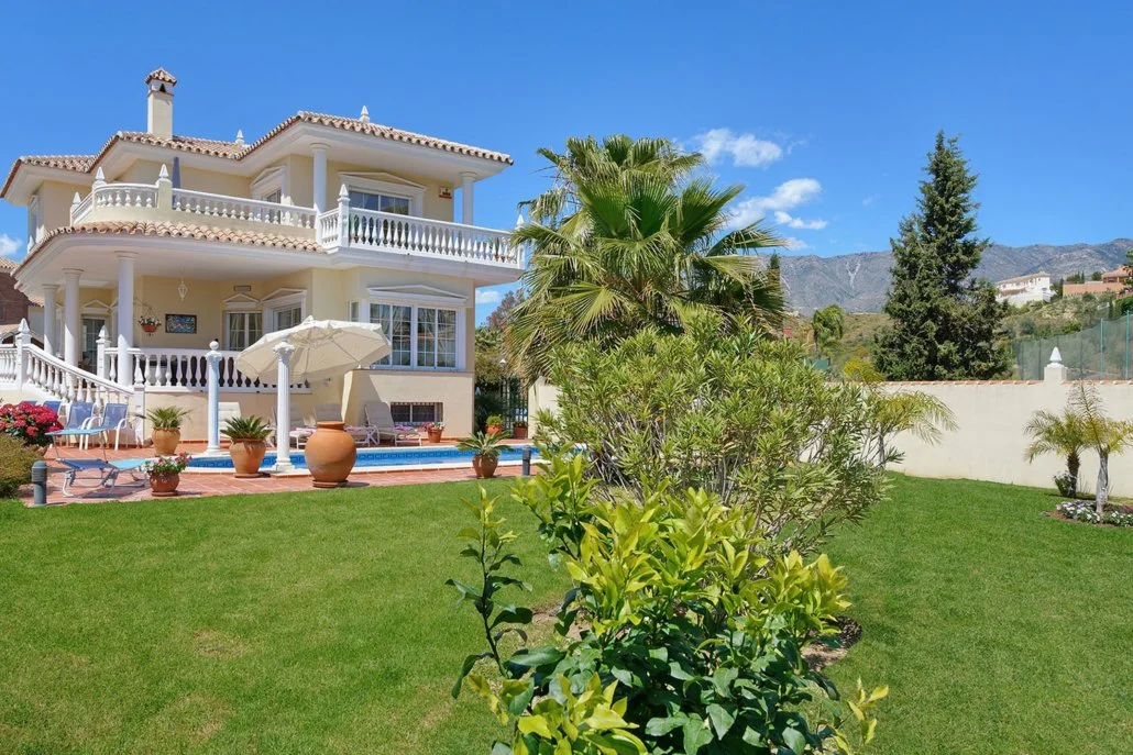 A large white two-story house with a tiled roof, multiple balconies, and a backyard with a swimming pool, patio furniture, potted plants, and palm trees. Mountainous landscape in the background under a clear blue sky.