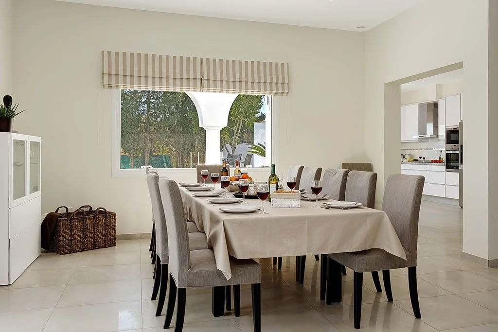 Dining room with a table set for dinner, including wine glasses, a bottle of wine, and plates. There is a window with a striped valance, and an open kitchen visible in the background.