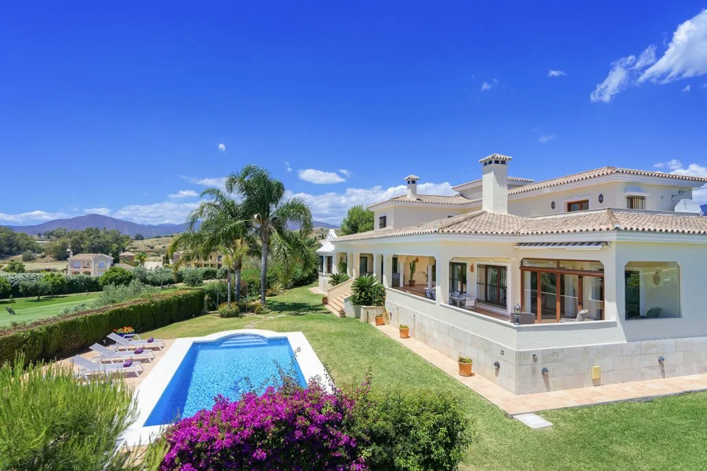 Large white house with a tiled roof, surrounded by a lush green lawn, palm trees, a swimming pool, and colorful flowers on a sunny day.