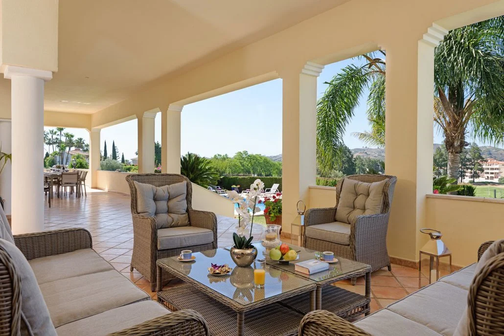 Porch with wicker chairs and cushioned seating, a glass-topped table with fruit, flowers, coffee cups, and sunlight streaming through open arches, overlooking a lush, green landscape with palm trees.