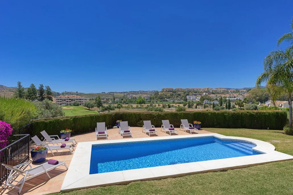 Swimming pool in a backyard with lounge chairs and potted plants, overlooking a hillside landscape under a clear blue sky.