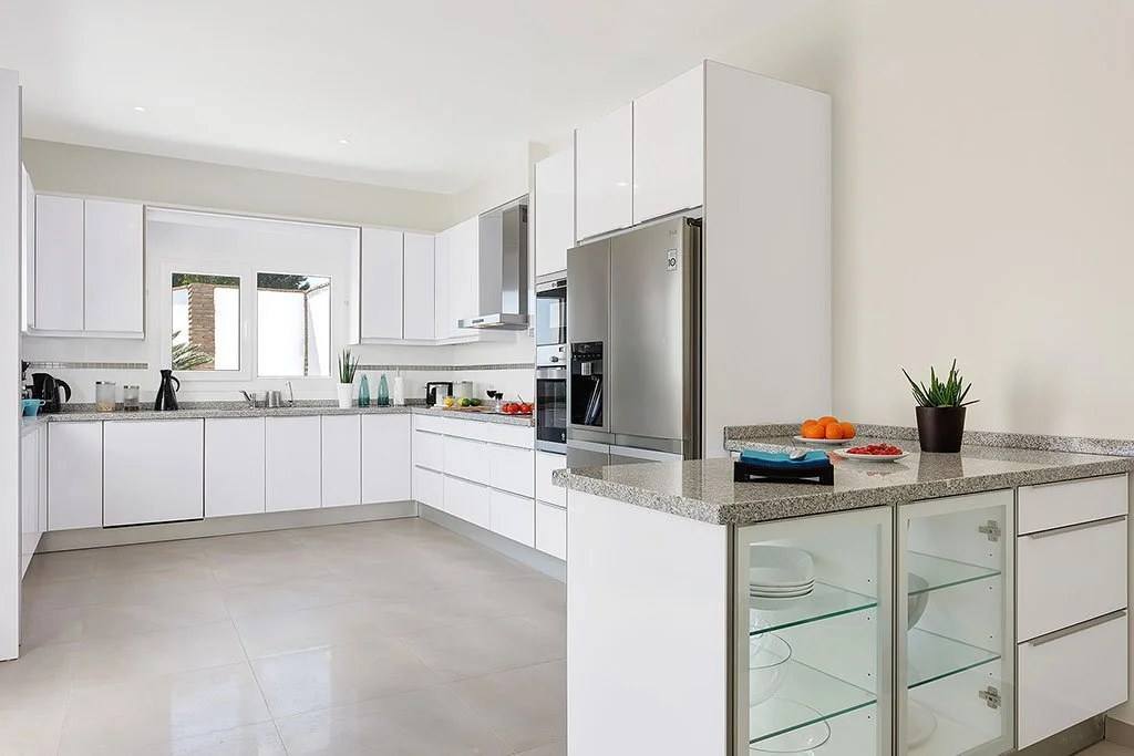 Modern white kitchen with granite countertops, stainless steel refrigerator, and minimal decor, including a potted plant and plates in glass display cabinets.