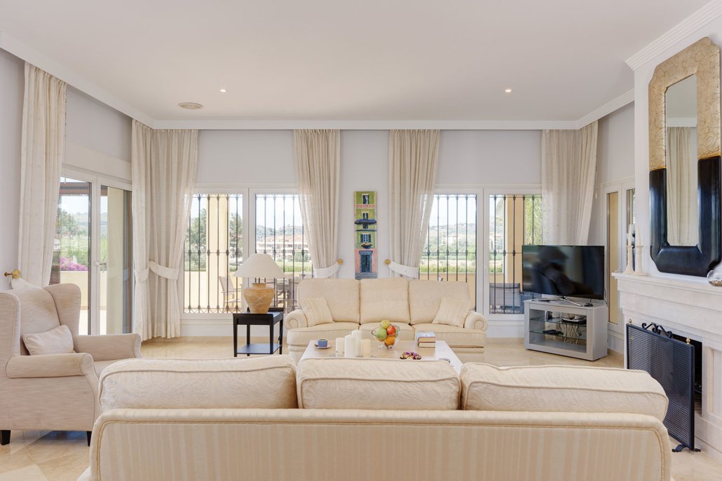 A bright, spacious living room with white sofas, a large flat-screen TV, and big windows with cream curtains.