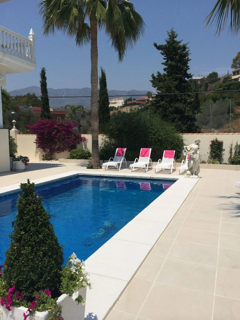 A backyard swimming pool area with three white lounge chairs with pink towels, a stone fountain sculpture, palm trees, flowering bushes, and a view of distant hills and houses under a clear blue sky.