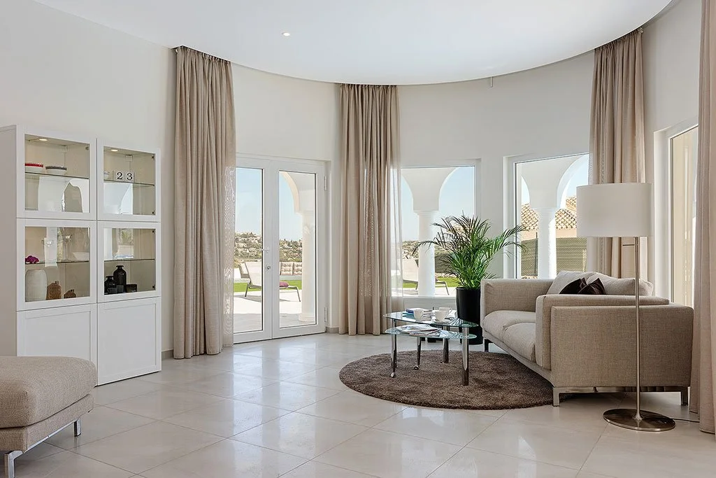 Bright, modern living room with large windows, beige curtains, a gray sofa, a round brown rug, a glass coffee table, a tall white floor lamp, and a potted plant.