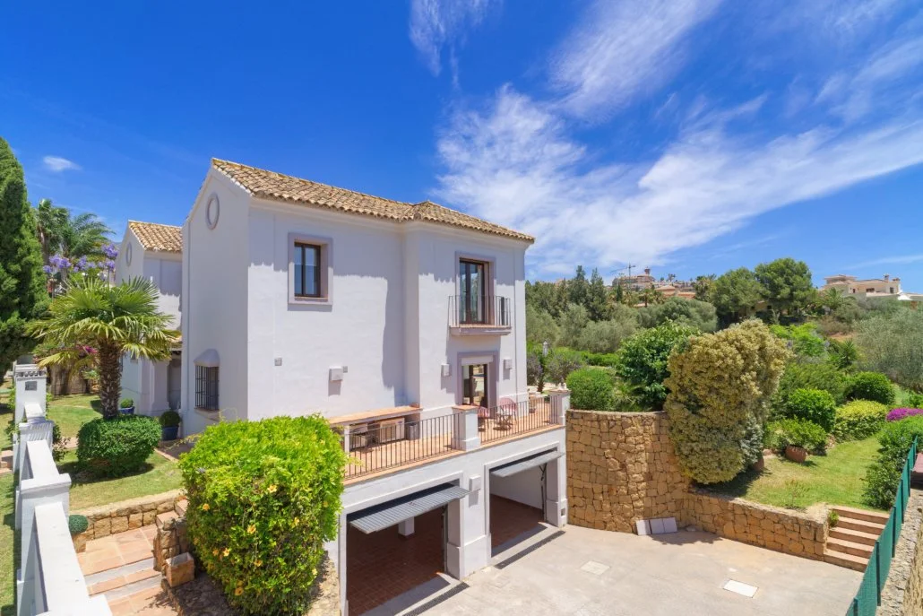 A white two-story house with a tiled roof, a balcony, and a garage, surrounded by trees and greenery under a blue sky with scattered clouds.