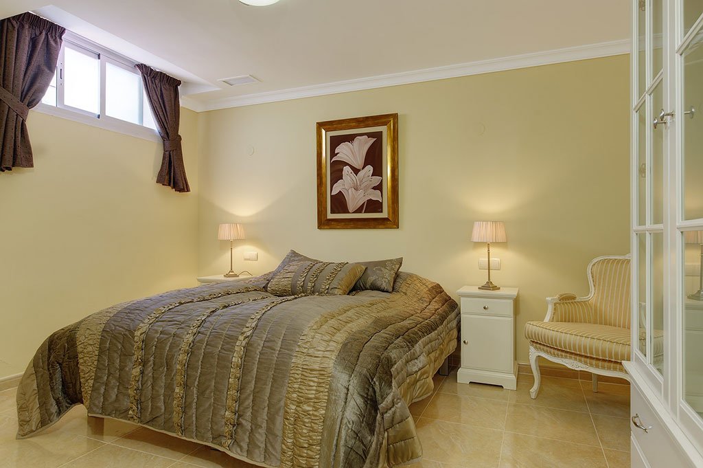 Bedroom with a large bed, two nightstands with lamps, striped armchair, framed floral artwork, small window with curtains, tiled floor.