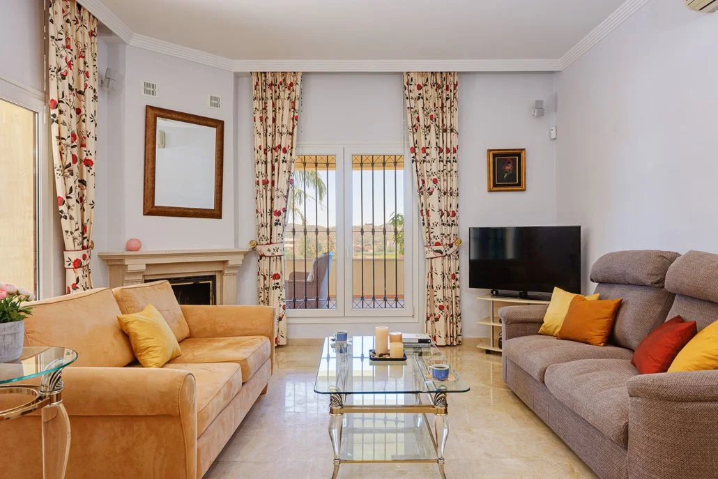 Living room with beige and gray sofas, colorful pillows, glass coffee table, fireplace, curtains, and a flat-screen TV.