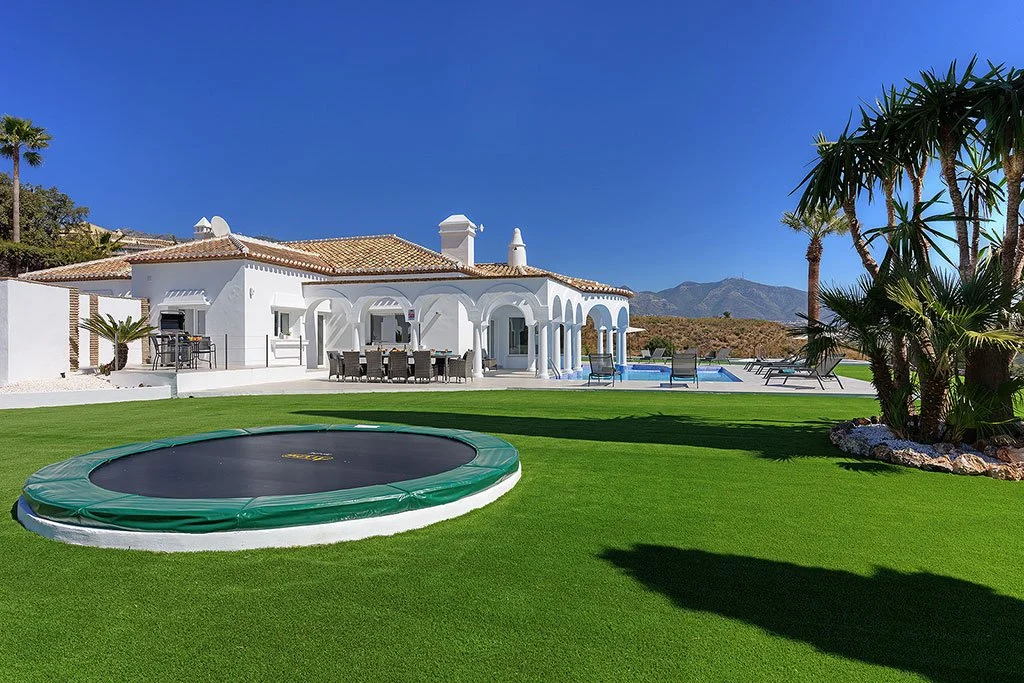 A backyard scene with green grass, a large trampoline, tropical plants, and a white house with a tiled roof, outdoor dining area, and a swimming pool with lounge chairs, set against a mountain backdrop under a clear blue sky.
