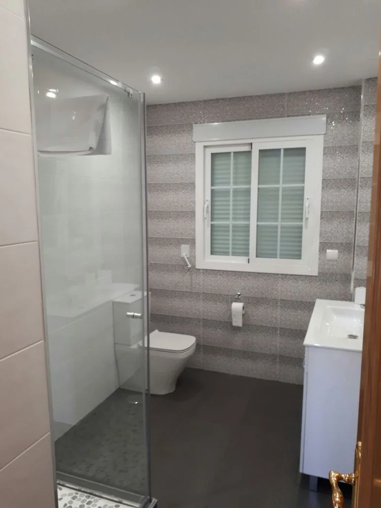 Modern bathroom with a glass shower enclosure, a white toilet, a window with shutters, and a white vanity with a sink.