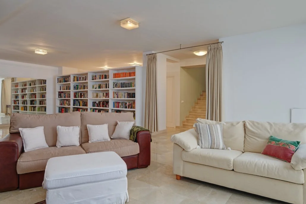 Living room with beige and white sofas, white bookshelf filled with books, and a staircase with wooden steps in the background.