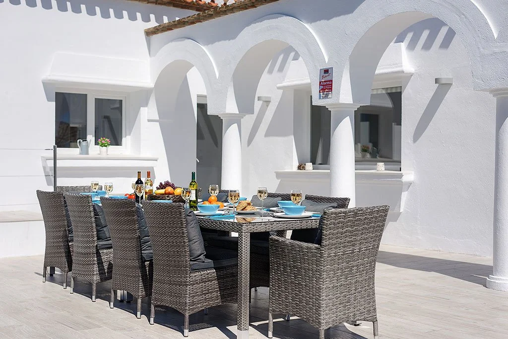 Outdoor dining setup on a patio with a table set for a meal, surrounded by wicker chairs, against a white Mediterranean-style building with arches.
