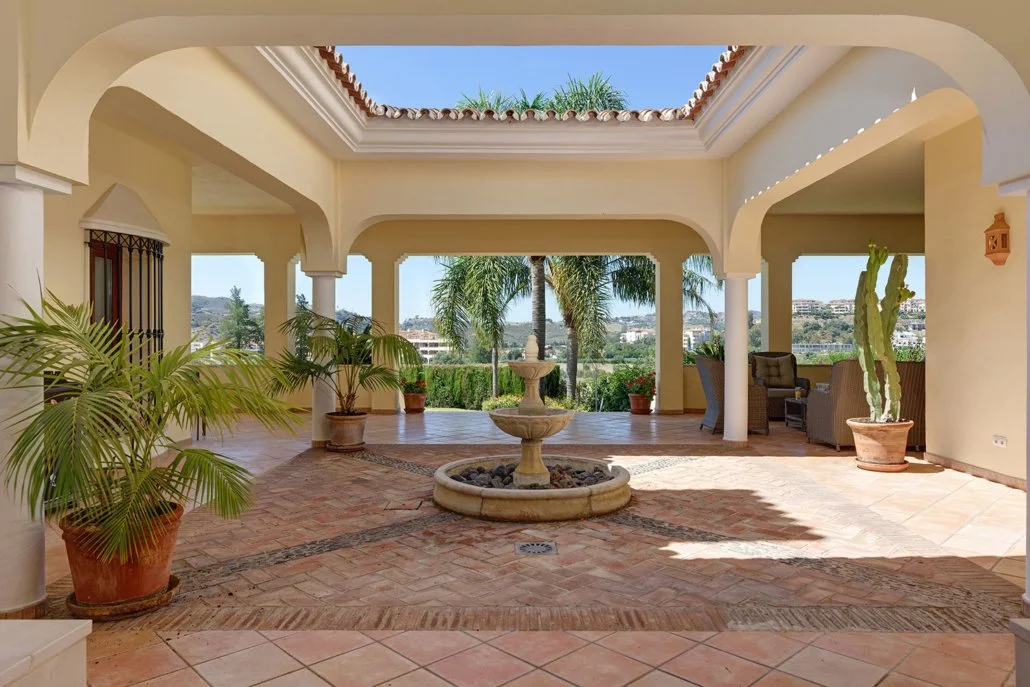A covered patio with a central fountain, surrounded by potted plants and outdoor seating, overlooking a landscape with trees and buildings in the distance.