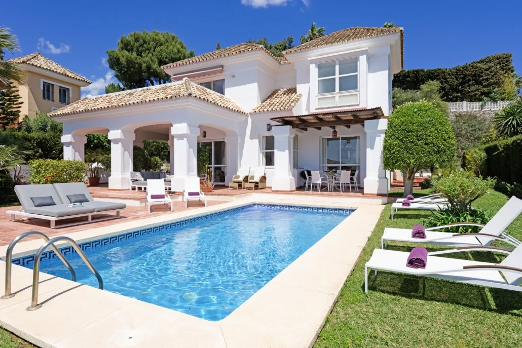 Exterior view of a white house with a tiled roof, swimming pool, lounge chairs, and lush greenery on sunny day.